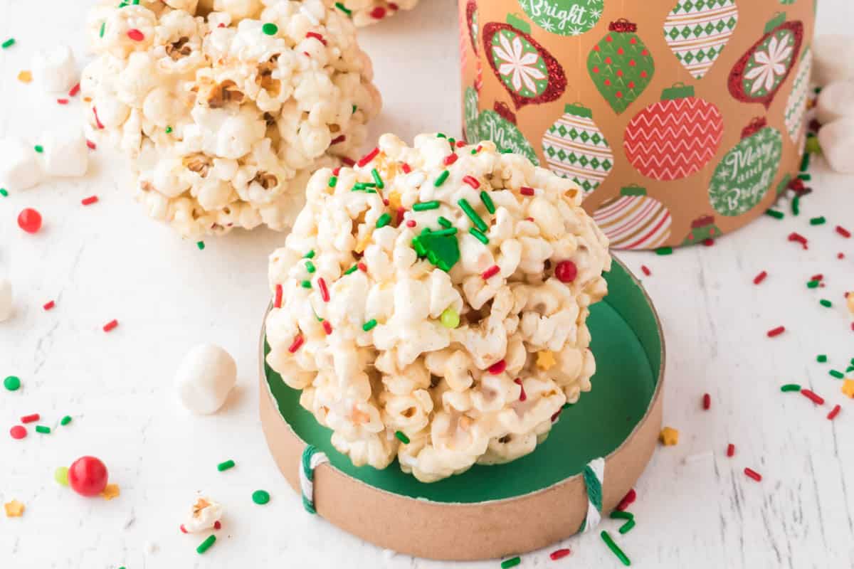 A popcorn ball covered with colorful holiday sprinkles sits on a round green lid, with a festive container and more popcorn balls in the background.