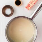 A saucepan with a creamy mixture next to a wooden spoon with vanilla, a dish of salt, and a stick of salted butter on a white surface.