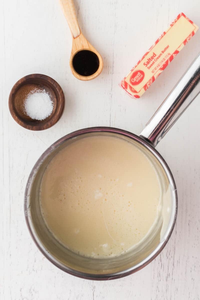 A saucepan with a creamy mixture next to a wooden spoon with vanilla, a dish of salt, and a stick of salted butter on a white surface.