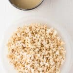 A bowl of plain popcorn sits on a white surface next to a metal bowl containing a light-colored sauce.