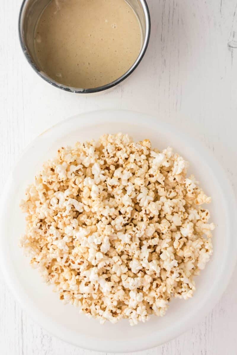 A bowl of plain popcorn sits on a white surface next to a metal bowl containing a light-colored sauce.