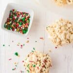 Two popcorn balls and a small bowl of red, green, and white holiday sprinkles on a white surface, with some sprinkles scattered nearby.