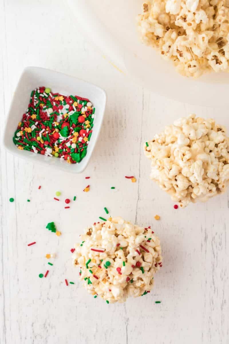 Two popcorn balls and a small bowl of red, green, and white holiday sprinkles on a white surface, with some sprinkles scattered nearby.