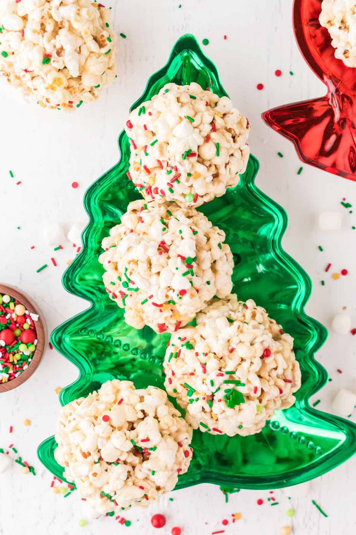 Five popcorn balls with red and green sprinkles are arranged on a green Christmas tree-shaped tray, surrounded by holiday-themed candies and sprinkles.