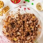A large glass bowl filled with chocolate-coated cereal mix sits on a white surface, surrounded by pretzels, red and green candies, and holiday decorations.