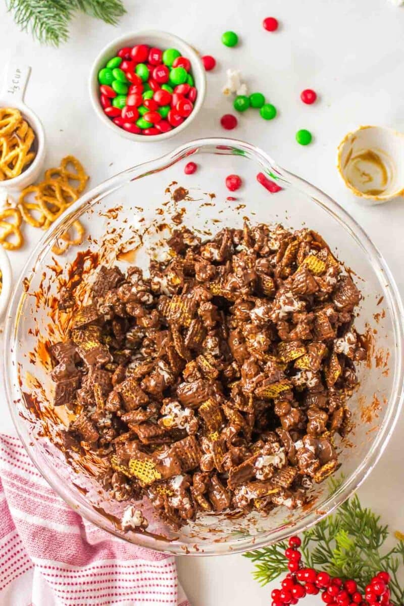 A large glass bowl filled with chocolate-coated cereal mix sits on a white surface, surrounded by pretzels, red and green candies, and holiday decorations.