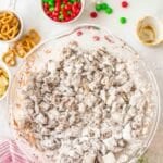 A glass bowl filled with powdered sugar-coated snack mix is on a countertop surrounded by pretzels, M&M’s, and holiday decorations.