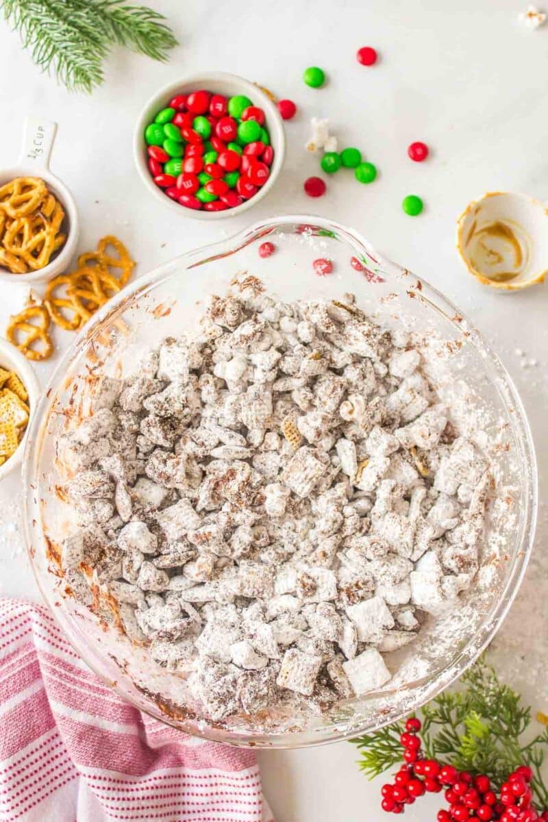 A glass bowl filled with powdered sugar-coated snack mix is on a countertop surrounded by pretzels, M&M&rsquo;s, and holiday decorations.