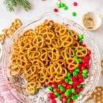 A glass bowl filled with pretzels, red and green candy-coated chocolates, and powdered sugar-coated cereal, surrounded by holiday decorations.