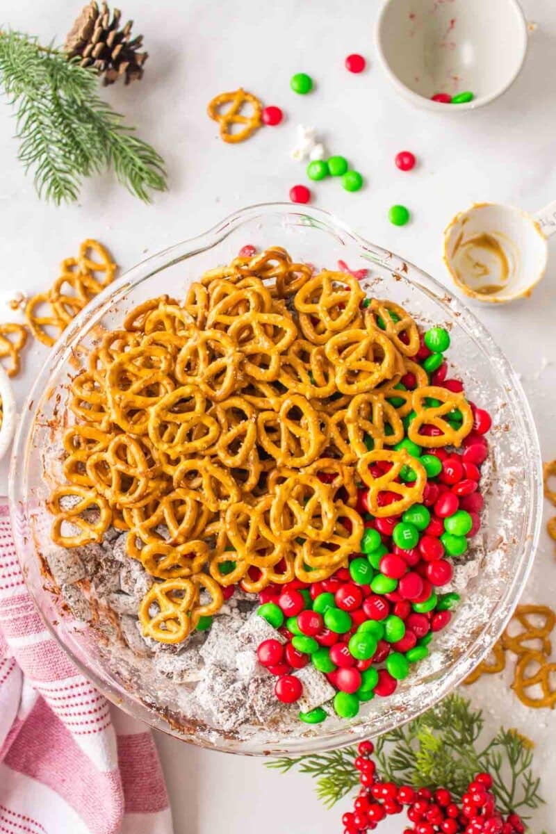 A glass bowl filled with pretzels, red and green candy-coated chocolates, and powdered sugar-coated cereal, surrounded by holiday decorations.