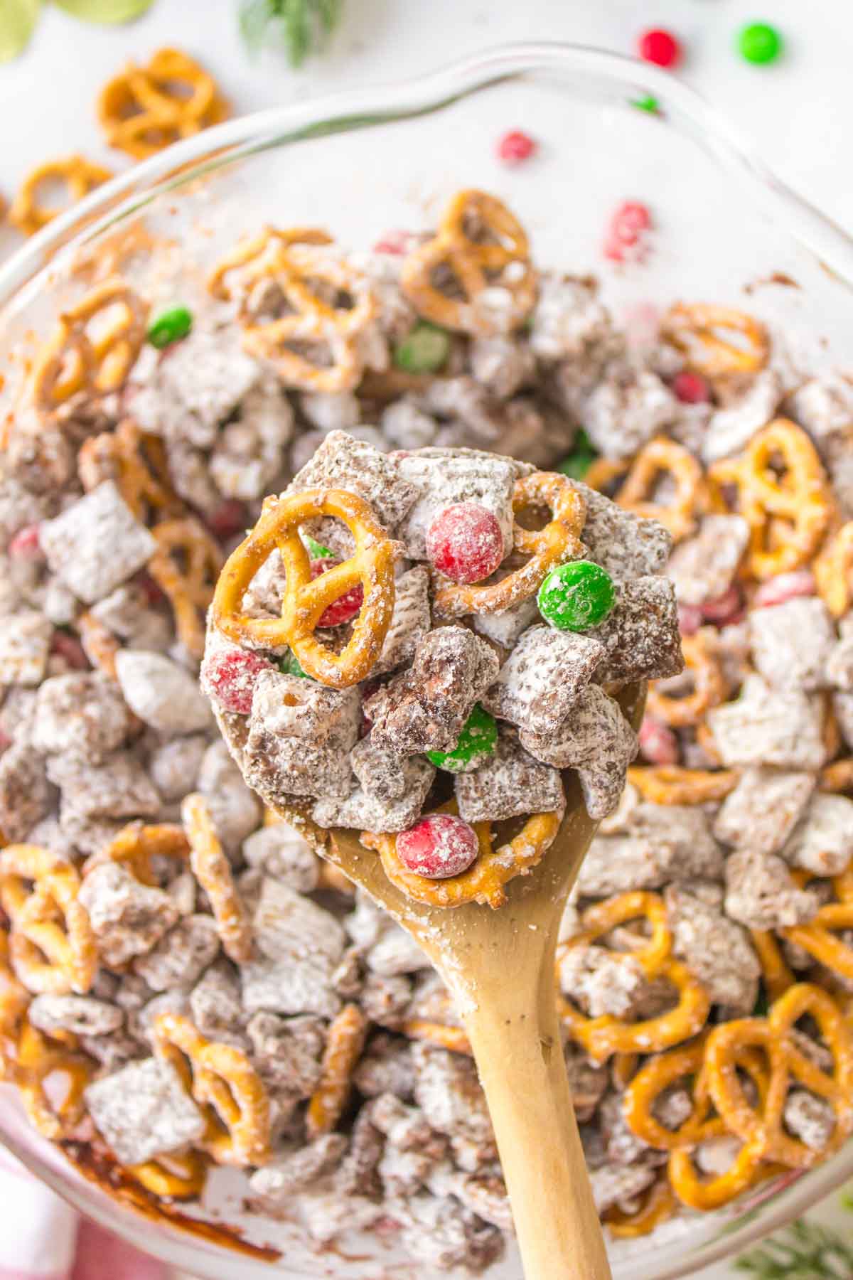 A wooden spoon holds a mix of pretzels, chocolate-coated cereal, powdered sugar, and red and green candy above a bowl of the same snack mix.