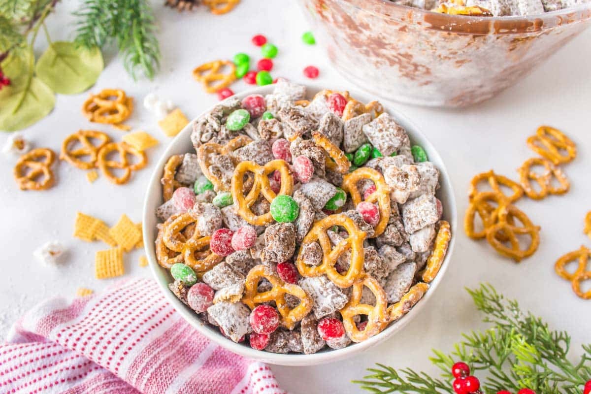 A bowl filled with powdered sugar-coated snack mix, pretzels, Chex cereal, and red and green candies, surrounded by scattered pretzels and holiday decorations.