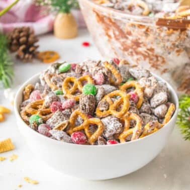 A white bowl filled with a snack mix of pretzels, Chex cereal, chocolate, powdered sugar, and red and green candies sits on a table with holiday decorations.