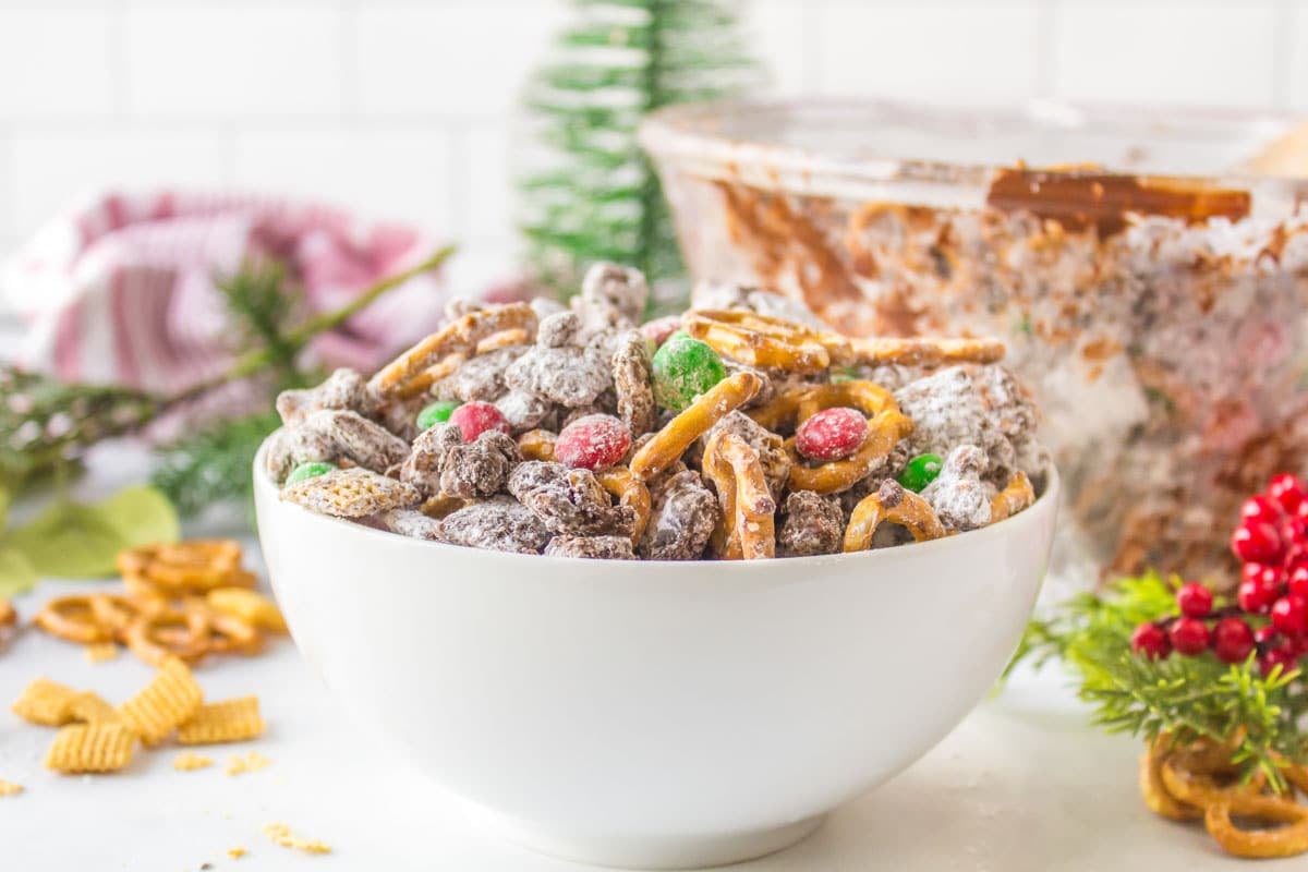 A white bowl filled with a snack mix of pretzels, Chex cereal, chocolate candies, and powdered sugar sits on a table with holiday decorations in the background.