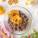 A glass bowl filled with chocolate chips and peanut butter, surrounded by pretzels, M&Ms, Chex cereal, and baking utensils on a white surface.