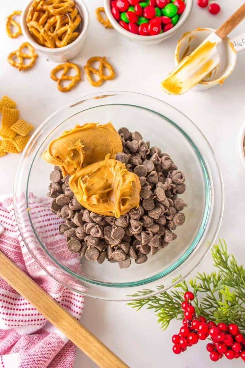 A glass bowl filled with chocolate chips and peanut butter, surrounded by pretzels, M&Ms, Chex cereal, and baking utensils on a white surface.