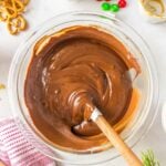A glass bowl of melted chocolate being stirred with a spatula, surrounded by pretzels, red and green M&Ms, and a striped kitchen towel.