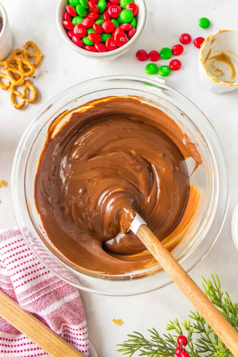 A glass bowl of melted chocolate being stirred with a spatula, surrounded by pretzels, red and green M&Ms, and a striped kitchen towel.