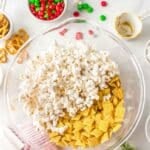 A glass mixing bowl containing popcorn and Chex cereal sits on a countertop, surrounded by pretzels, holiday M&Ms, measuring cups, and festive decorations.