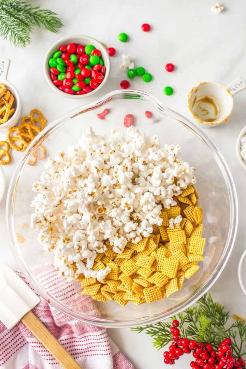 A glass mixing bowl containing popcorn and Chex cereal sits on a countertop, surrounded by pretzels, holiday M&Ms, measuring cups, and festive decorations.