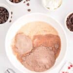 A mixing bowl with cake mix, eggs, and liquid ingredients surrounded by bowls of chocolate chips, sour cream, and an electric mixer on a white surface.