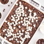 A rectangular baking dish filled with chocolate brownies topped with mini marshmallows, next to bowls of chocolate chips, frosting, and whipped cream.