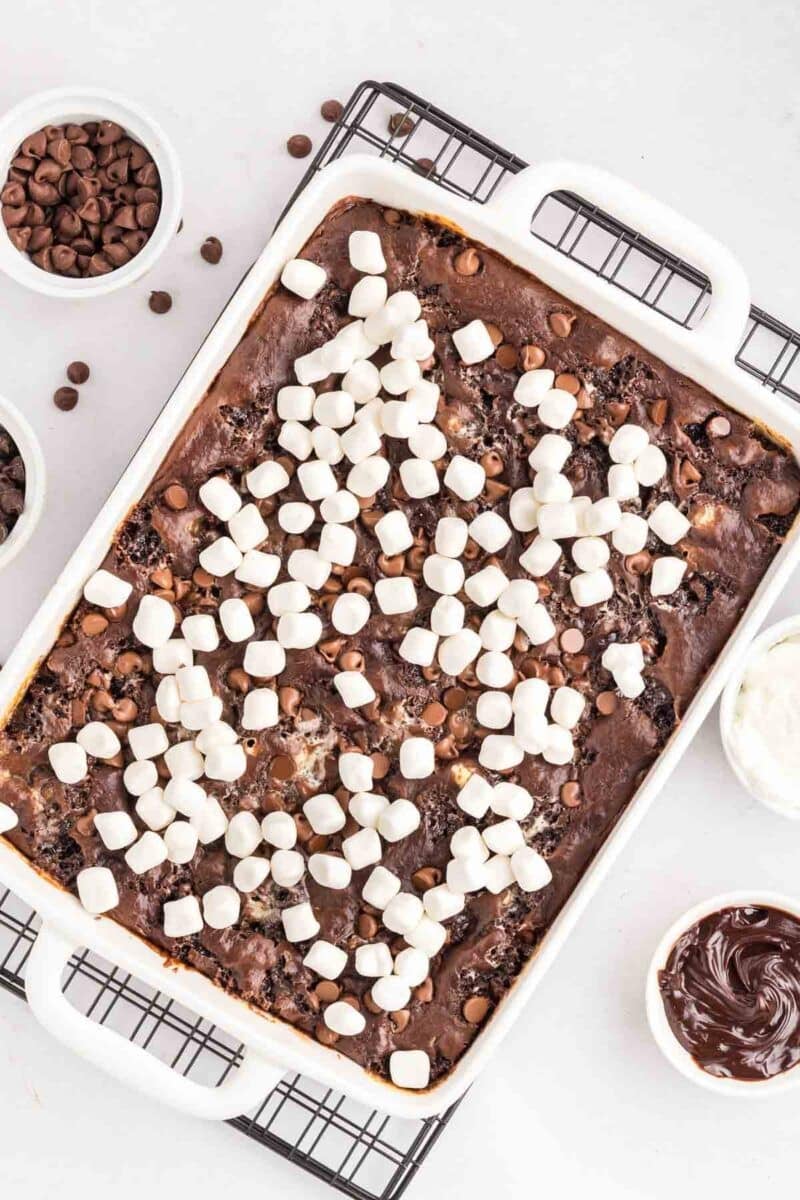 A rectangular baking dish filled with chocolate brownies topped with mini marshmallows, next to bowls of chocolate chips, frosting, and whipped cream.