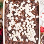A baked chocolate dessert in a white rectangular dish topped with mini marshmallows and chocolate chips, surrounded by Christmas-themed decorations.