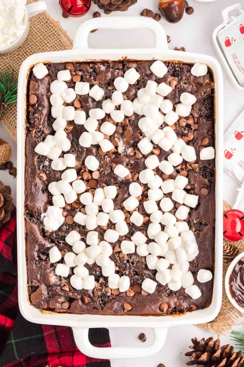 A baked chocolate dessert in a white rectangular dish topped with mini marshmallows and chocolate chips, surrounded by Christmas-themed decorations.