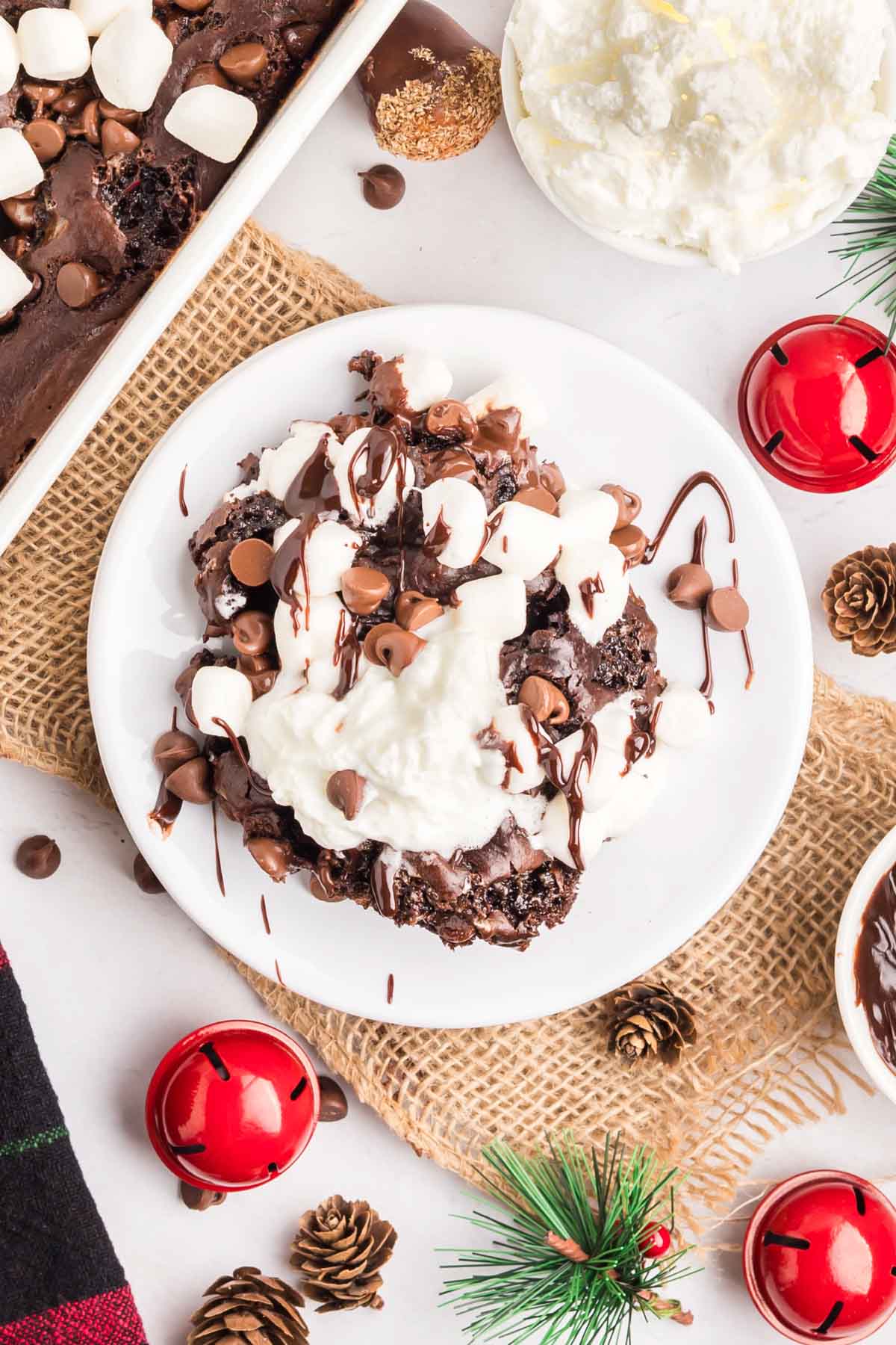 A plate of chocolate brownies topped with whipped cream, marshmallows, and chocolate chips, surrounded by Christmas decorations.