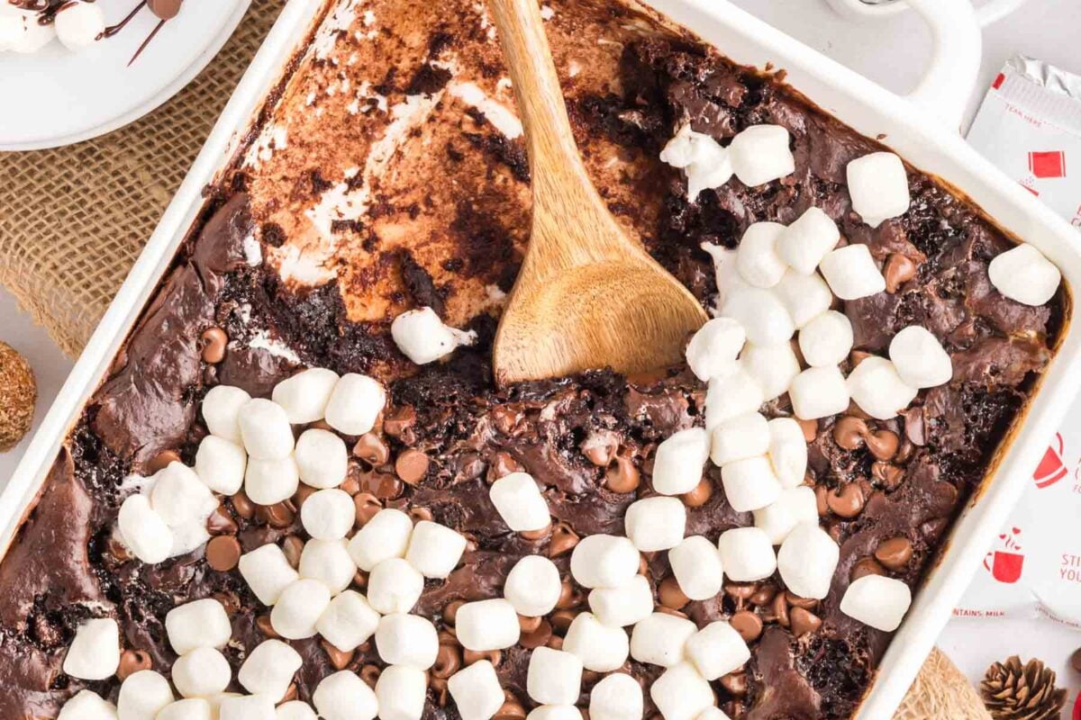 A baked chocolate dessert topped with mini marshmallows and chocolate chips in a white dish, with a wooden spoon resting inside.