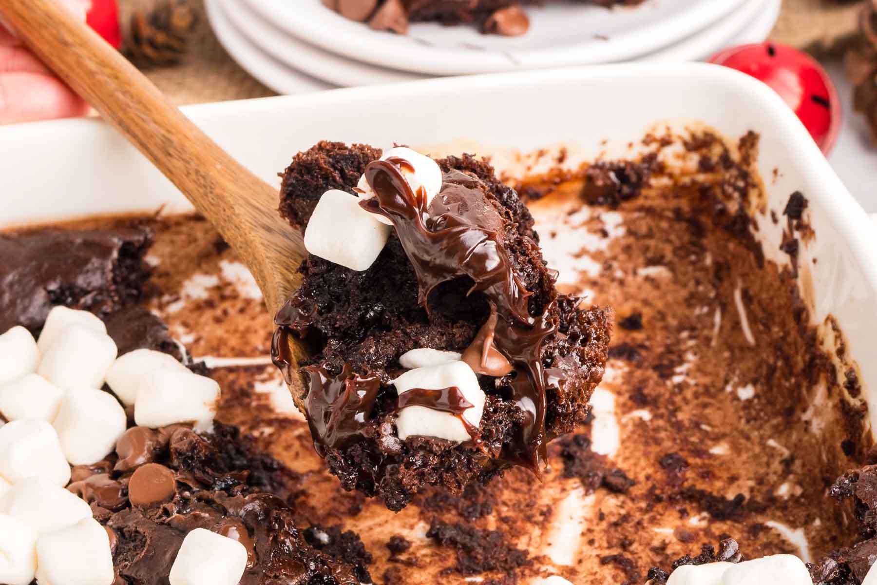 A wooden spoon lifts a serving of chocolate brownies topped with melted chocolate and mini marshmallows from a baking dish.