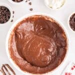 A bowl of chocolate cake batter surrounded by chocolate chips, sour cream, a hand mixer with chocolate on the beaters, and baking ingredient packets on a white surface.