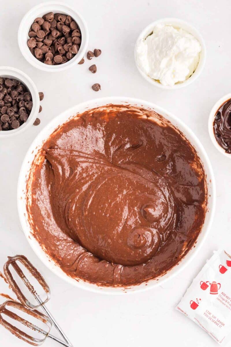 A bowl of chocolate cake batter surrounded by chocolate chips, sour cream, a hand mixer with chocolate on the beaters, and baking ingredient packets on a white surface.