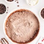 A mixing bowl with dry chocolate cake mix, surrounded by chocolate chips, whipped topping, electric mixer beaters, and packets on a white surface.