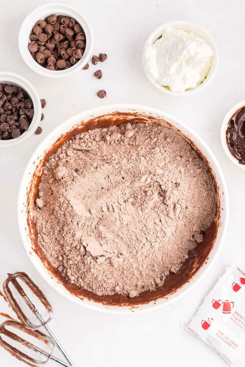 A mixing bowl with dry chocolate cake mix, surrounded by chocolate chips, whipped topping, electric mixer beaters, and packets on a white surface.