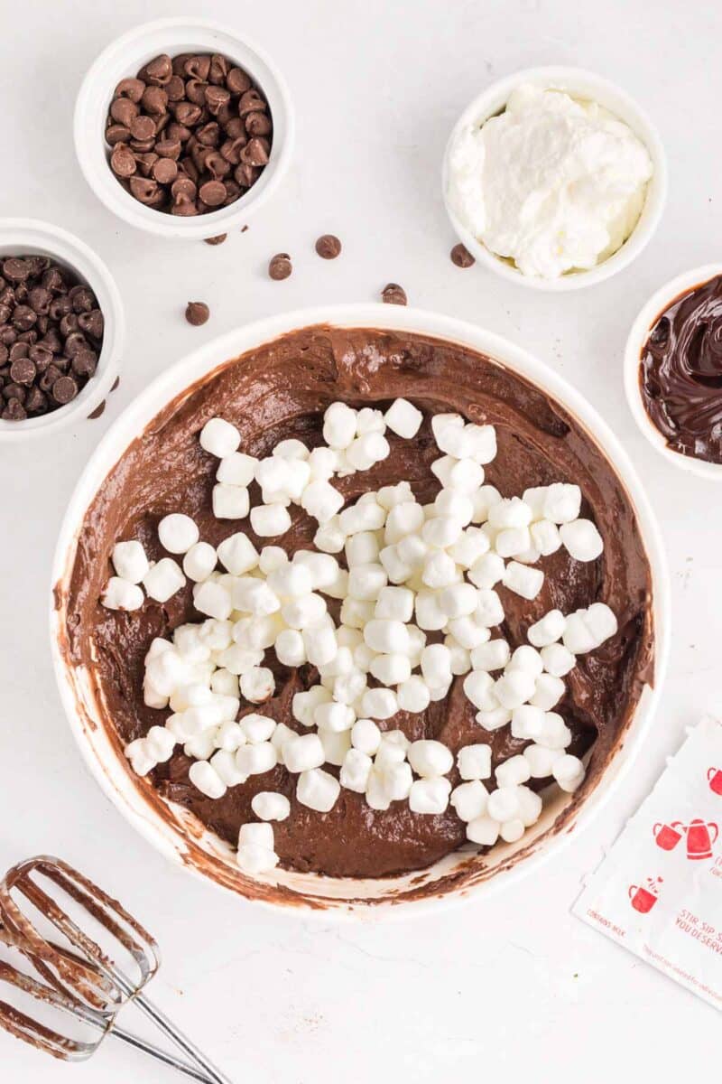 A bowl of chocolate batter topped with mini marshmallows, surrounded by bowls of chocolate chips, whipped topping, and chocolate spread, with a mixer and a packet nearby.