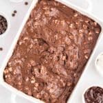 A white baking dish filled with chocolate brownie batter, surrounded by bowls of chocolate chips, chocolate sauce, and cream on a white surface.