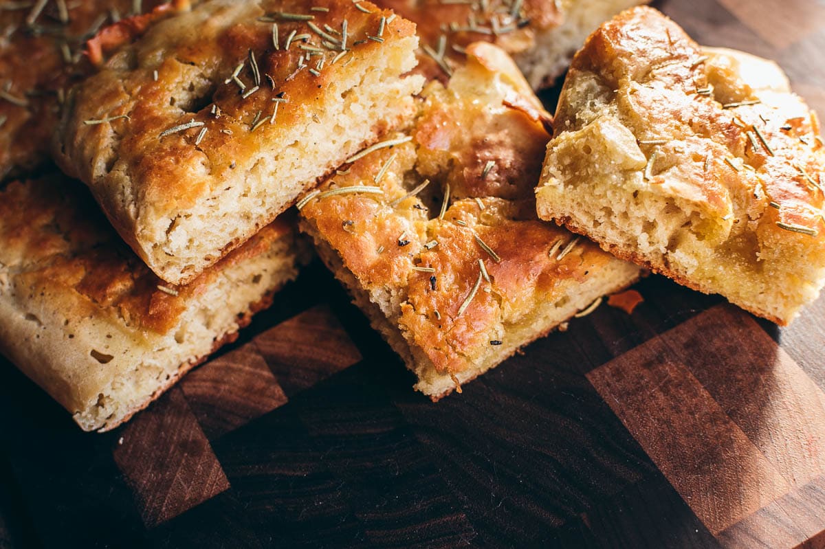 Sliced pieces of golden-brown focaccia bread with rosemary on a dark wooden cutting board—perfect for joyful vegan dining or as part of your festive vegan recipes.