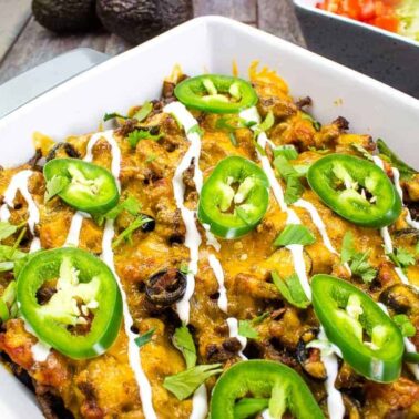 A casserole dish filled with baked Mexican-style beef casserole, topped with melted cheese, sliced jalapeños, green onions, and drizzled with sour cream. Its rich layers and bold appeal are complemented by a bowl of lettuce and tomatoes in the background.