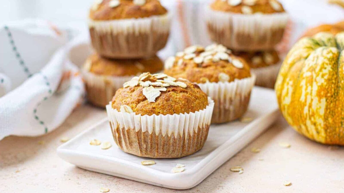Pumpkin muffins topped with oats—a true pantry staple—are arranged on a white rectangular plate, with more muffins and a pumpkin in the background, making them perfect for anyone seeking tasty oat recipes.