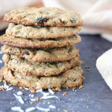 A stack of six oatmeal cookies with visible oats and raisins sits on a dark surface, surrounded by cookie crumbs and coconut flakes—perfect as satisfying snacks or energizing bites for any time of day.