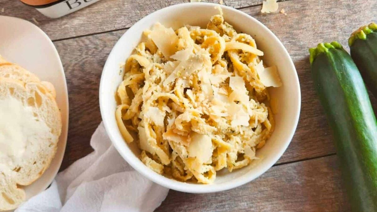 A white bowl filled with pasta topped with shaved cheese sits on a wooden surface next to two zucchinis, a plate of sliced bread, and a white napkin—perfect inspiration for new zucchini recipes.