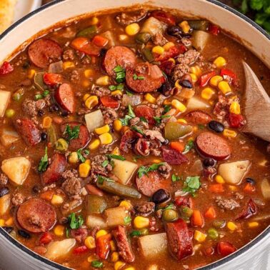 A pot of hearty stew with sausage, ground beef, potatoes, beans, corn, carrots, tomatoes, and green beans—one of those cozy meals and easy beef recipes—served with a wooden spoon and cornbread on the side.
