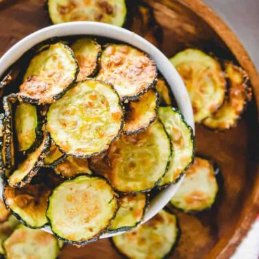 A bowl of baked zucchini chips, a tasty addition to your favorite zucchini recipes, sits inside a larger wooden bowl filled with more chips, next to a white and red striped cloth.