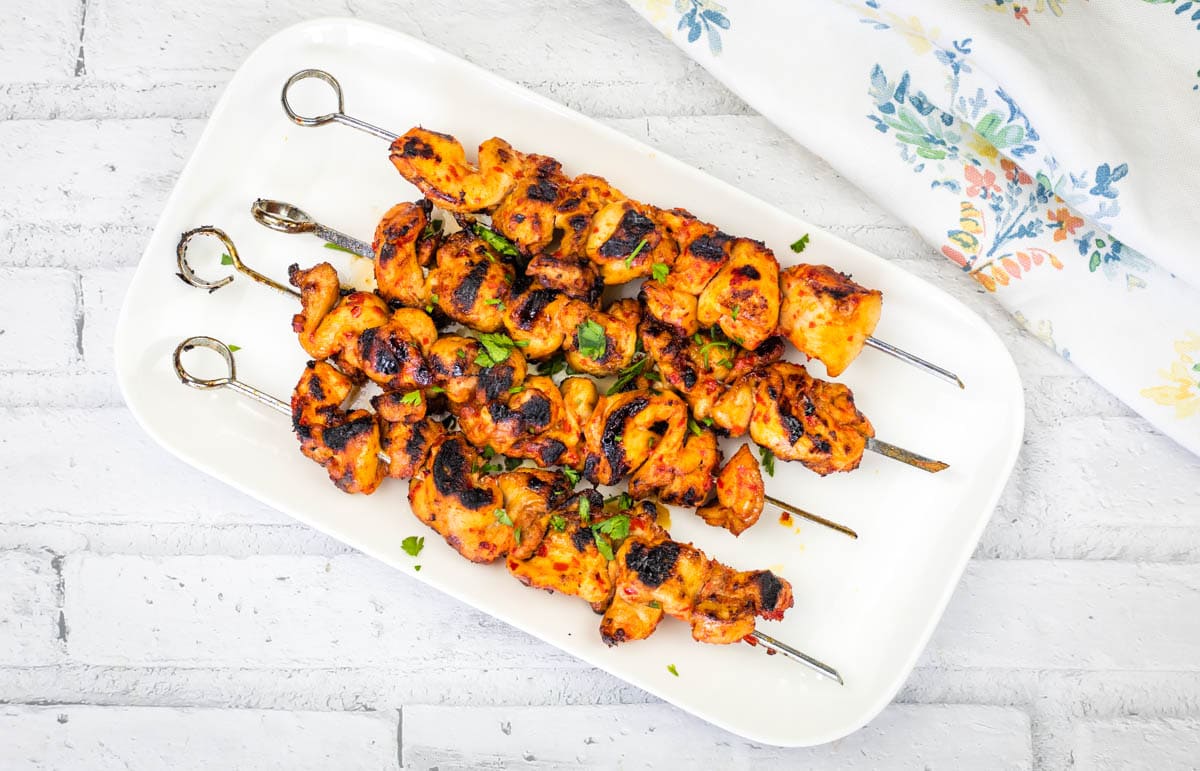 Four grilled chicken skewers, perfect for effortless dinner ideas and grilled recipes, are garnished with chopped herbs and arranged on a white rectangular plate, set on a light brick background beside a floral-patterned napkin.