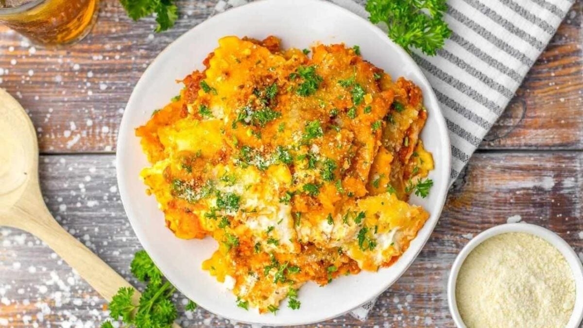 A plate of baked lasagna topped with melted cheese and fresh parsley sits on a wooden table next to a bowl of grated cheese and a striped napkin, making it one of those classic comfort recipes perfect for filling meals.