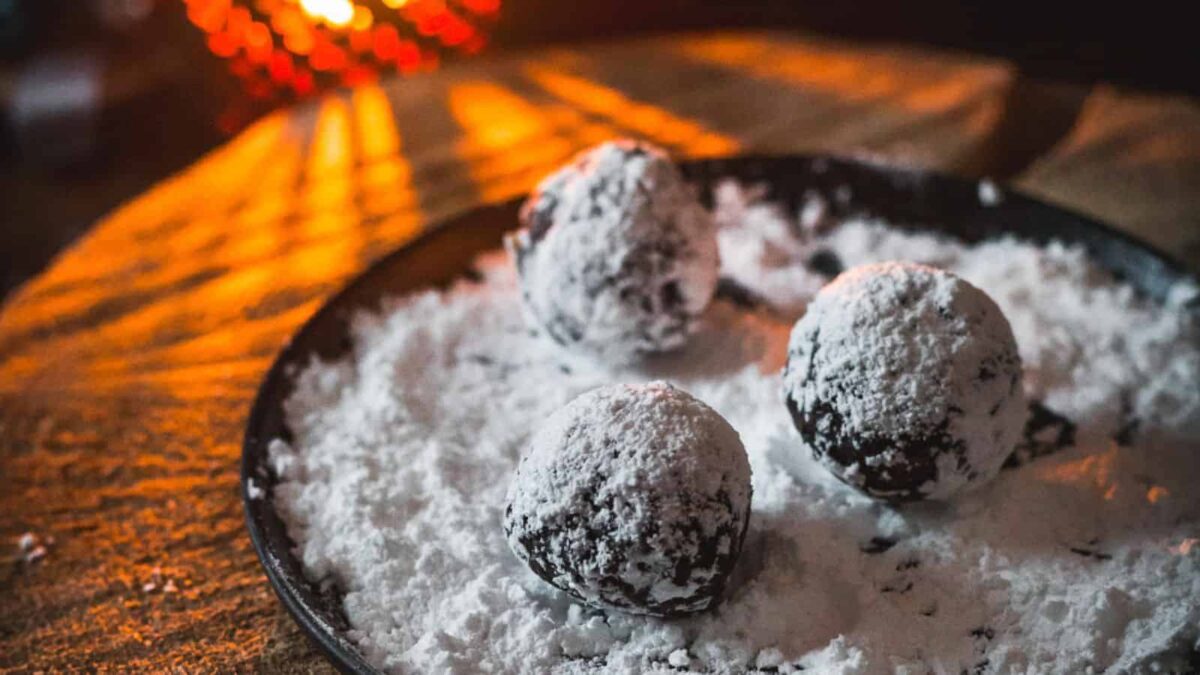Three festive chocolate truffles coated in powdered sugar rest on a plate dusted with more powdered sugar, with warm lighting in the background—perfect for vegan dining or holiday Christmas recipes.