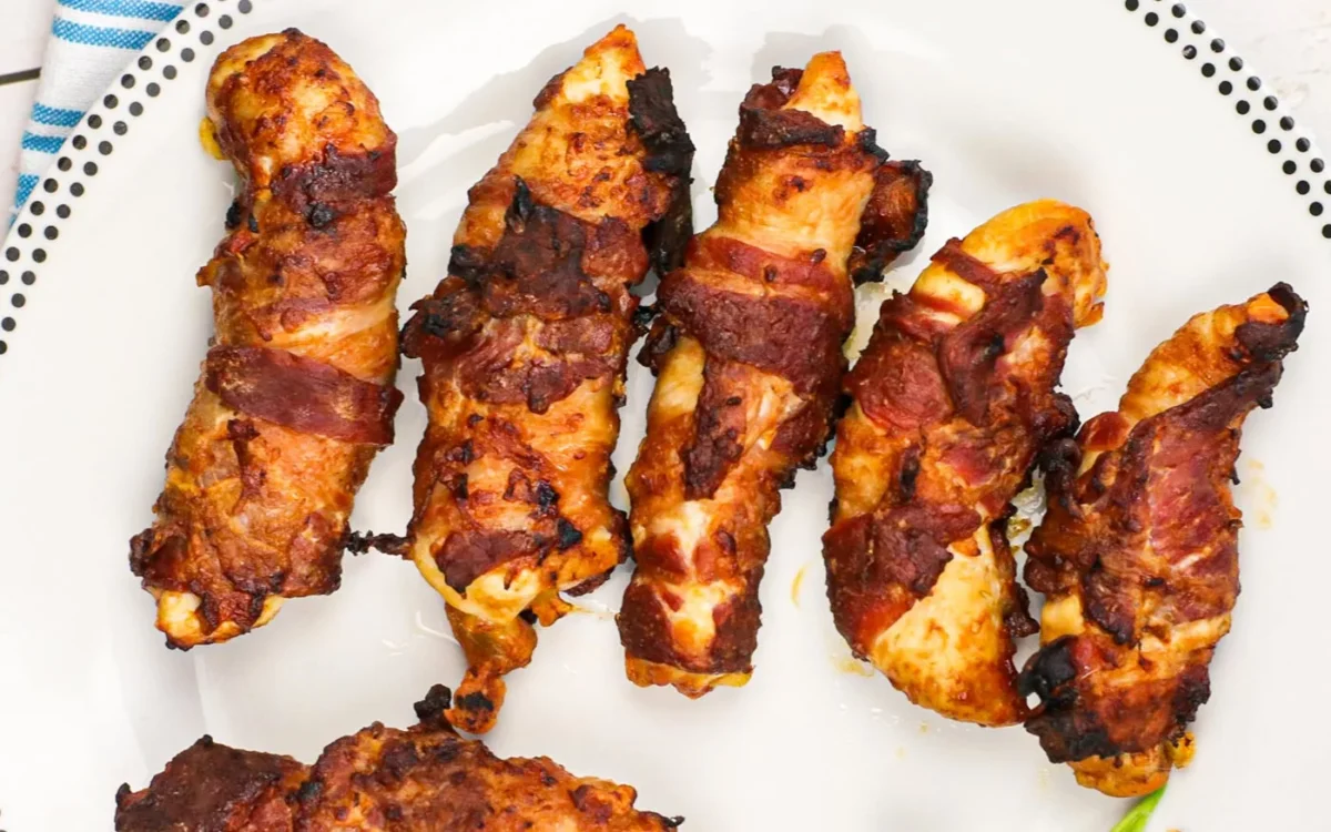 Five crispy, bacon-wrapped chicken tenders on a white plate with a patterned rim.