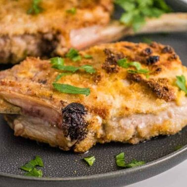 A breaded and cooked pork chop with a bone is served on a dark plate, garnished with chopped parsley—perfect for Everyday Cooking and Quick and Easy dinners.